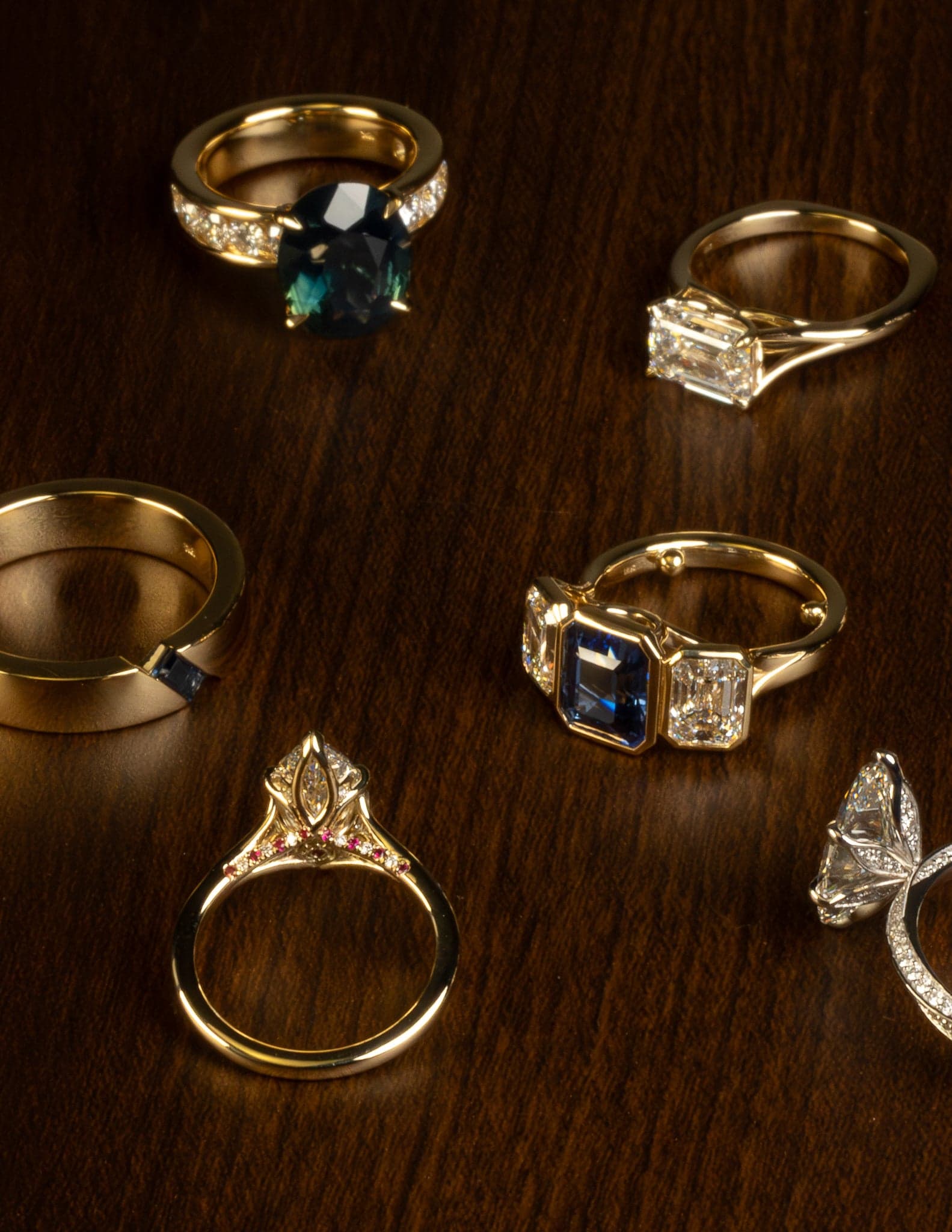 Nine Minhè commissions — yellow and white gold, sapphire, marquise and emerald cuts — arranged on a walnut bench.
